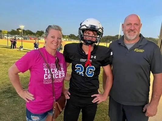 A football player in uniform with helmet stands between two adults. They are outside on a field.