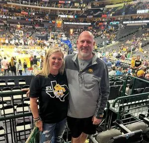 Two people at a basketball game. Woman in black shirt, man in gray jacket, both smiling, stadium in the background.