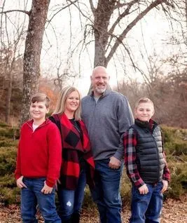 Family of four poses outdoors; two boys, a woman in buffalo plaid, and a man, standing near trees.
