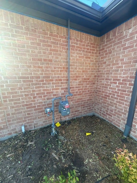 Gas meter and piping installation against a brick wall, with dark mulch and some green plants on the ground.
