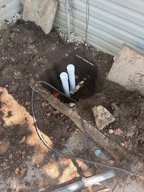 An excavated trench containing two white PVC pipes connecting to a smaller white PVC pipe, surrounded by dirt and debris.