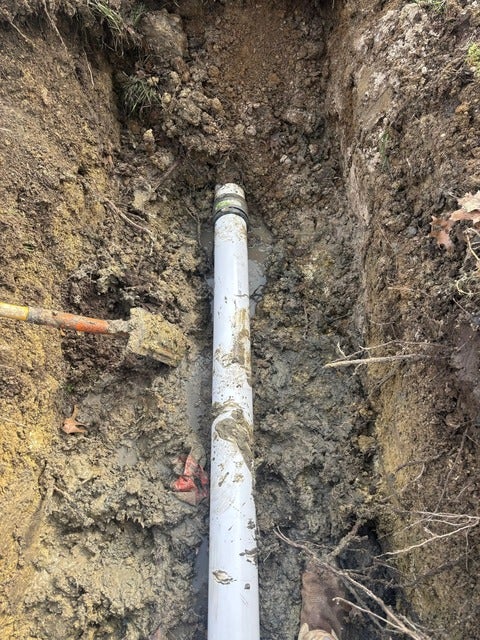 A white PVC pipe running vertically through a deep, muddy trench with a shovel visible on the left.