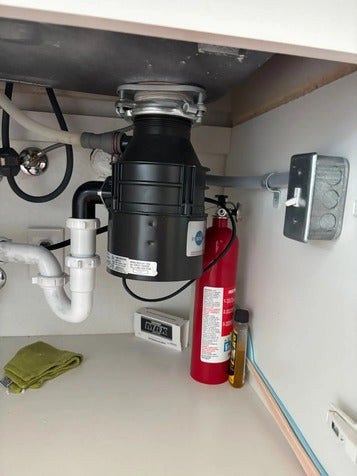 Under-sink cabinet with a garbage disposal, plumbing, a fire extinguisher, a light switch, and a washcloth.