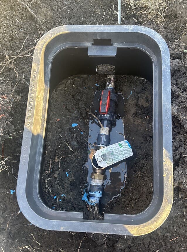 A buried irrigation valve box contains a water meter assembly with a partially submerged valve, water, and muddy dirt.