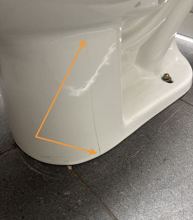 A vertical crack on the white base of a toilet, highlighted by an orange arrow emphasizing the crack's length.