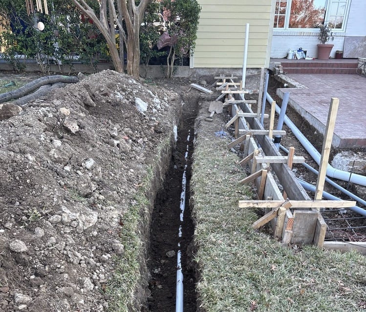 A long trench with newly installed white pipes, a dirt pile, and wooden concrete forms beside a house.