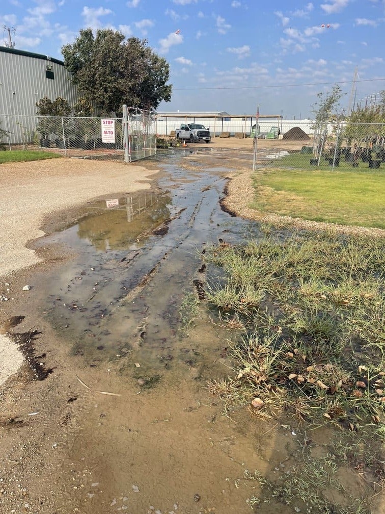 A muddy, water-filled track with tire ruts in front of a chain-link fence, leading to a commercial lot.