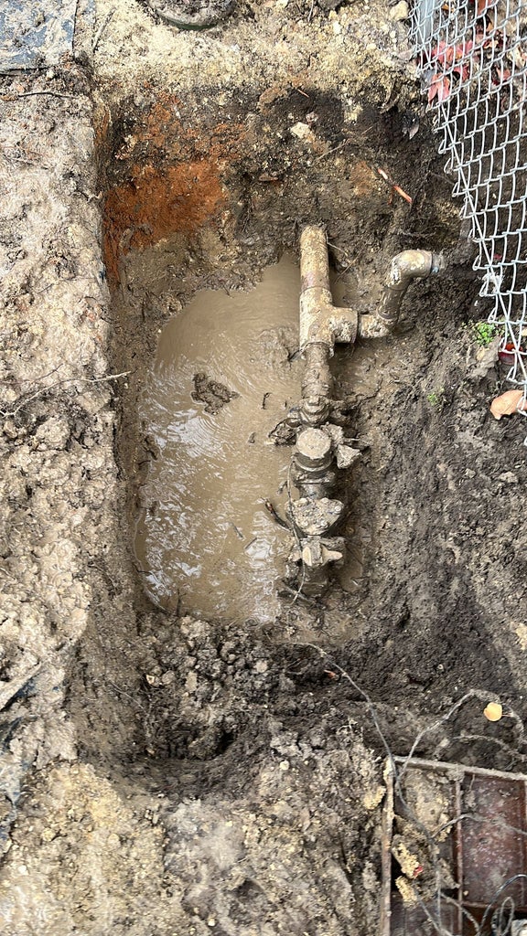 A deep, narrow trench filled with muddy water, exposing a corroded pipe with valves and connections.