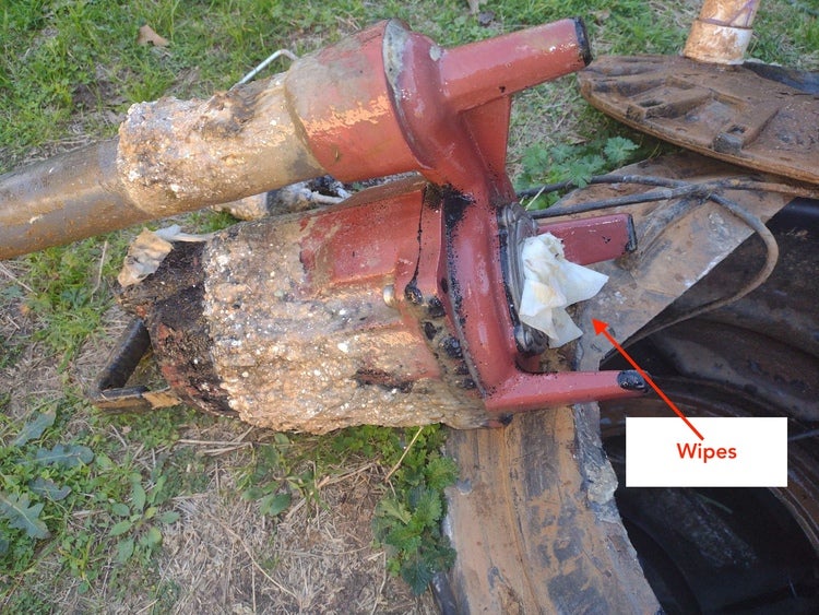 A red wastewater pump heavily corroded with white solids and wrapped in white wipes, sitting on grass next to an open manhole.