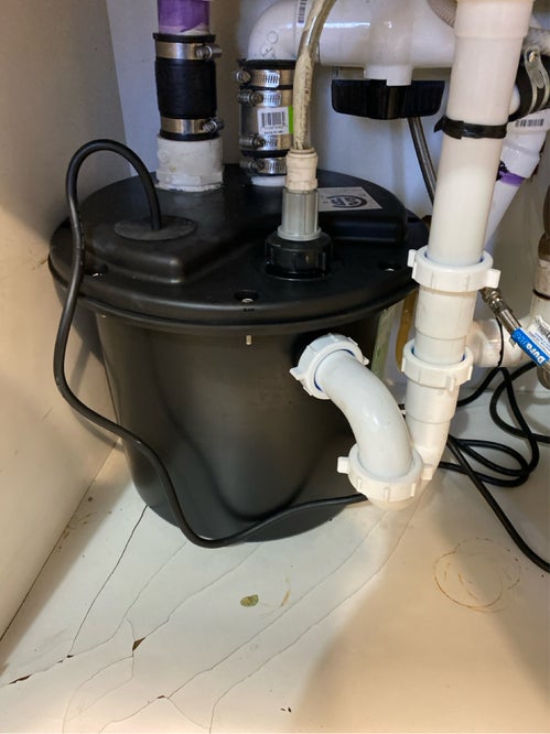 Dark gray sewage grinder pump with multiple pipes, wires, and a white P-trap connected underneath a sink.