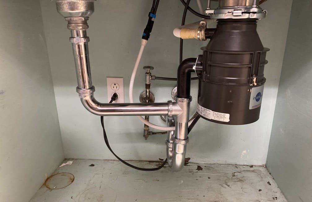 Kitchen sink disposal and P-trap plumbing under a counter, against an aqua wall with an electrical outlet.
