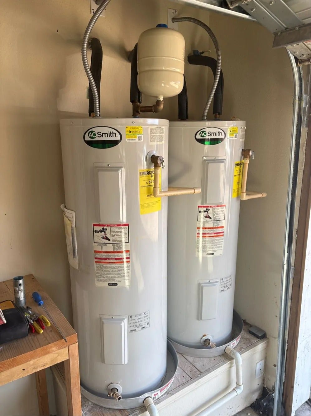 Two large, white A.O. Smith water heaters with yellow energy guide labels sit in a garage corner.