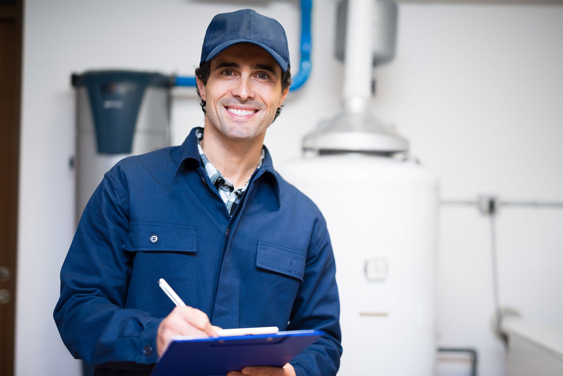 A smiling male plumber in a blue uniform and cap writes on a clipboard with a boiler in the background.