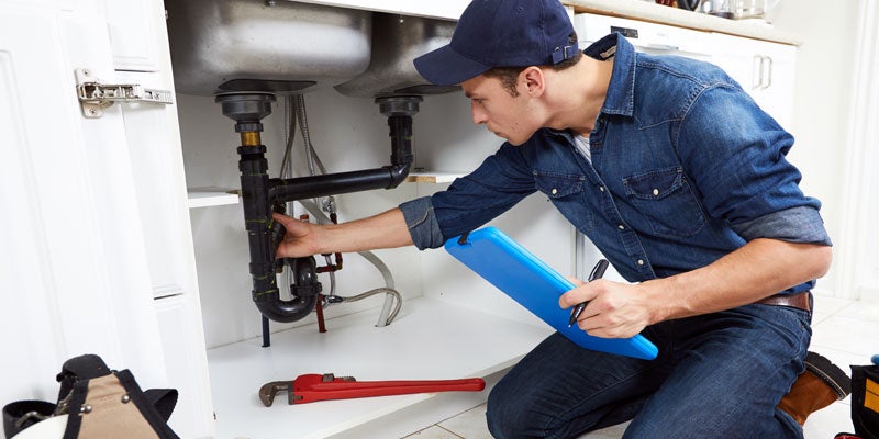 A plumber in a baseball cap and denim shirt, kneeling and inspecting pipes under a kitchen sink.