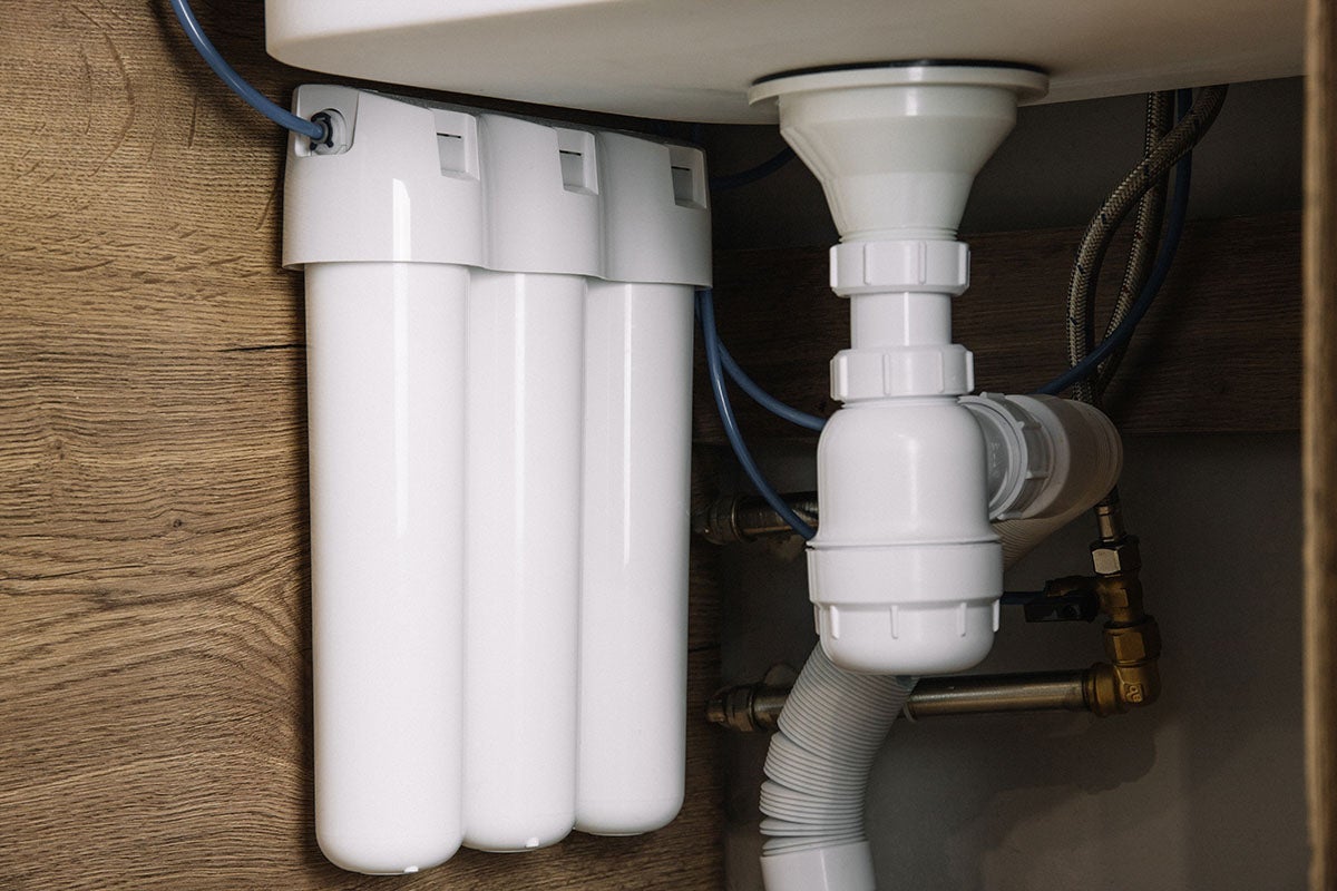 Under-sink water filter system with three white cartridges, next to a white plastic sink drain and plumbing.