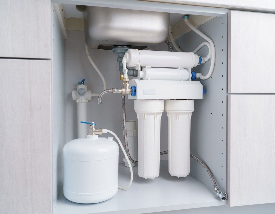 White water filtration system with tank, filters, and tubing installed under a kitchen sink in a light gray cabinet.
