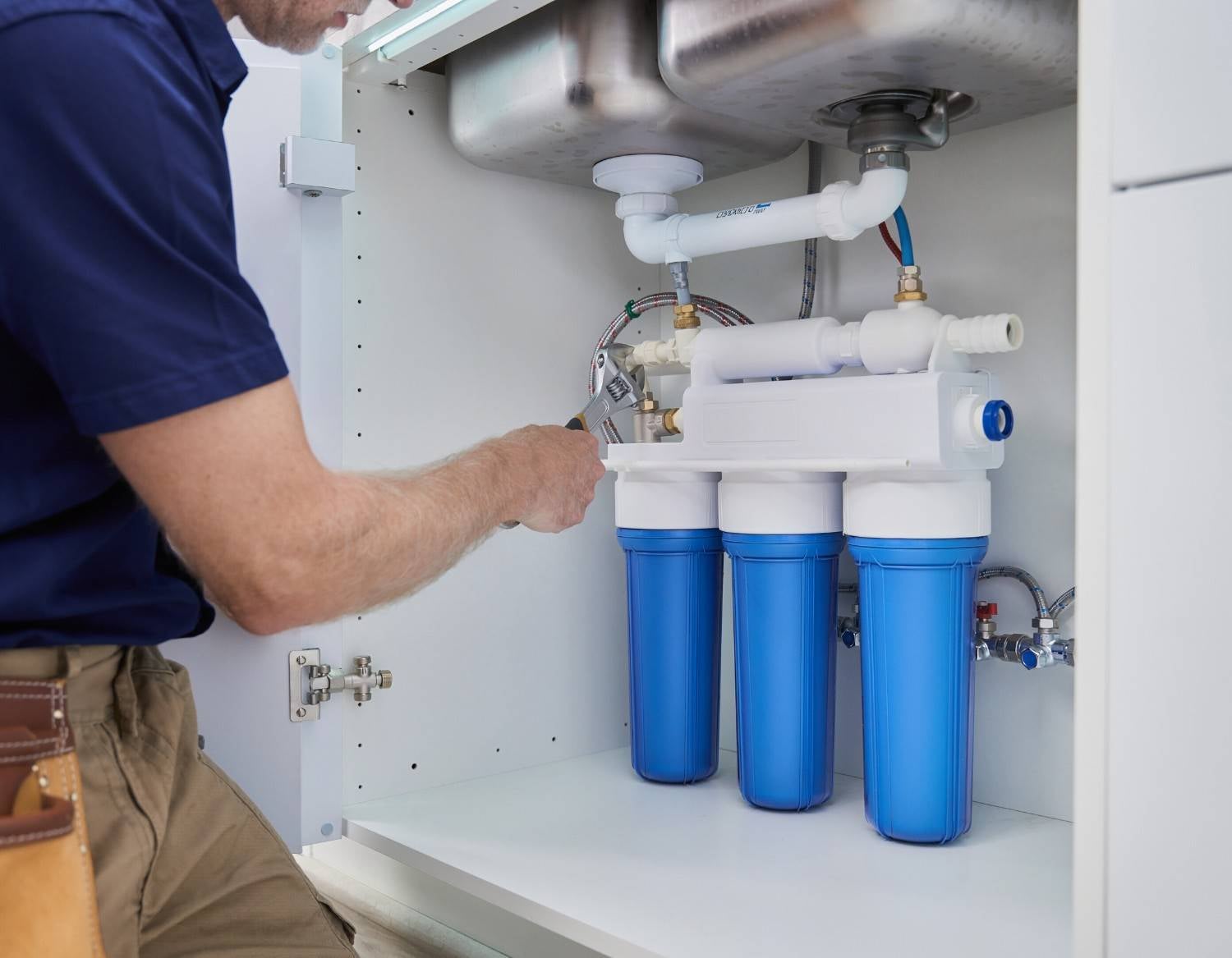 Plumber installing a triple-stage water filter system with three blue cartridges under a kitchen sink.